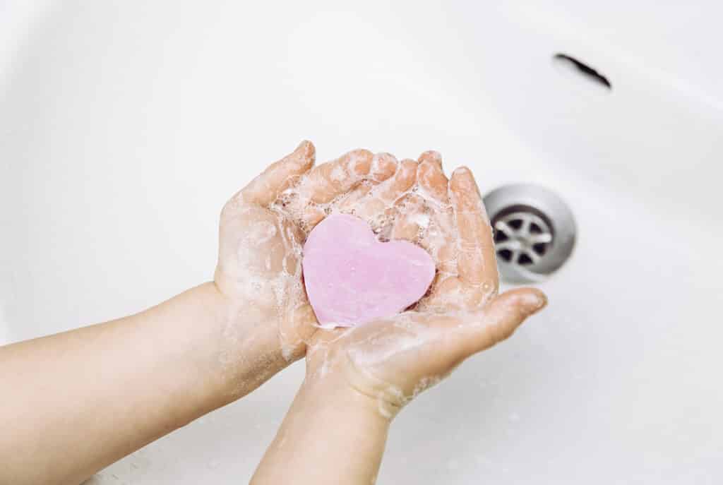 Importance of personal hygiene care. Flat lay view of child washing dirty hands with pink heart shape soap bar, lot of foam. Copy space.